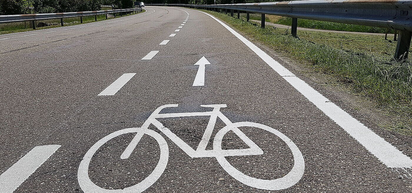 Eine Landstraße auf der ein Schutzstreifen für Radfahrende mit einer weißen gestrichelten Linie markiert ist.