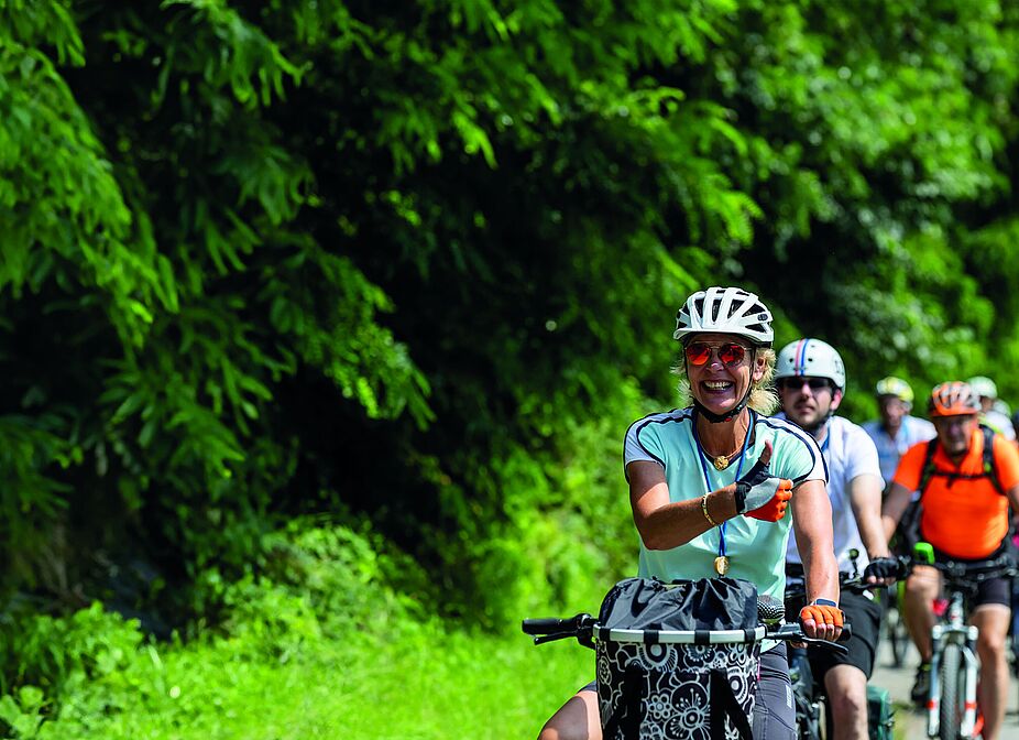 NRW Radtour 2025 Eine Gruppe von Radfahrenden auf einer Straße entlang eines Waldes.
