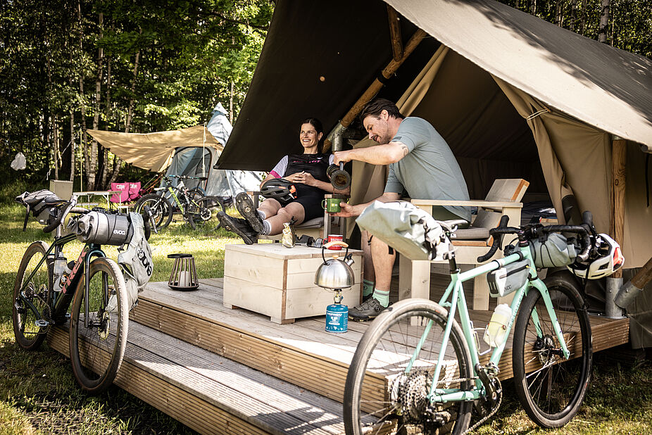 Bett+Bike Sport Unterkunft für Gravelbike Zwei Menschen mit Gravelbikes sitzen im Eingangsbereich auf einer Holzveranda vor einem großen Zelt.