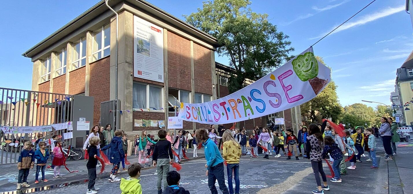 Schulstraße Kinder verschiedenen Alters spielen und bewegen sich auf einer gepflasterten Straße vor einem zweistöckigen Backsteingebäude. Über der Straße hängt ein buntes Banner mit der Aufschrift „Schulstraße“ und einem grünen Baum-Symbol. Im Hintergrund sind Erwachsene zu sehen, die die Aktion begleiten.