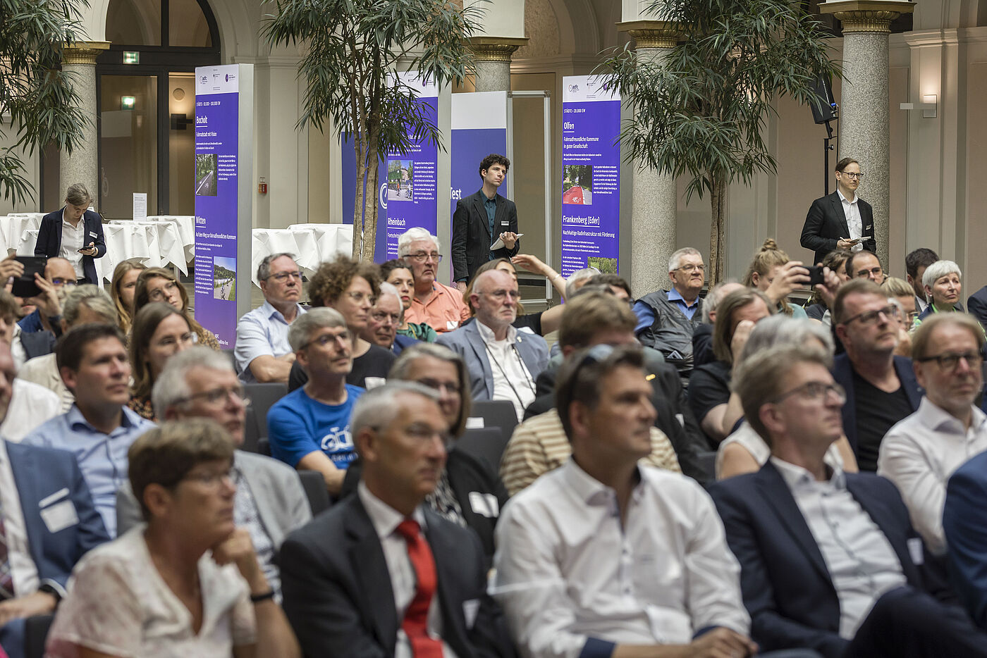 Großer Veranstaltungssaal mit vielen Besuchenden in Reihen, überwiegend Männer und Frauen mittleren Alters in Business-Kleidung, im Hintergrund lila Banner zum ADFC-Fahrradklima-Test, Palmen und Säulen in repräsentativem Gebäude, Fotografen dokumentieren die Veranstaltung