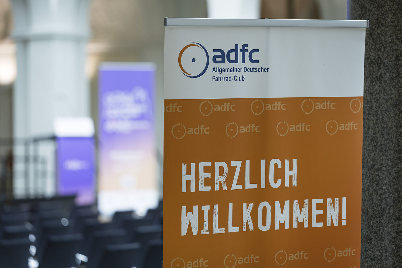 Oranges ADFC-Banner mit weißer Schrift "HERZLICH WILLKOMMEN!" und ADFC-Logo, im Hintergrund unscharf lila Banner und Veranstaltungsbereich